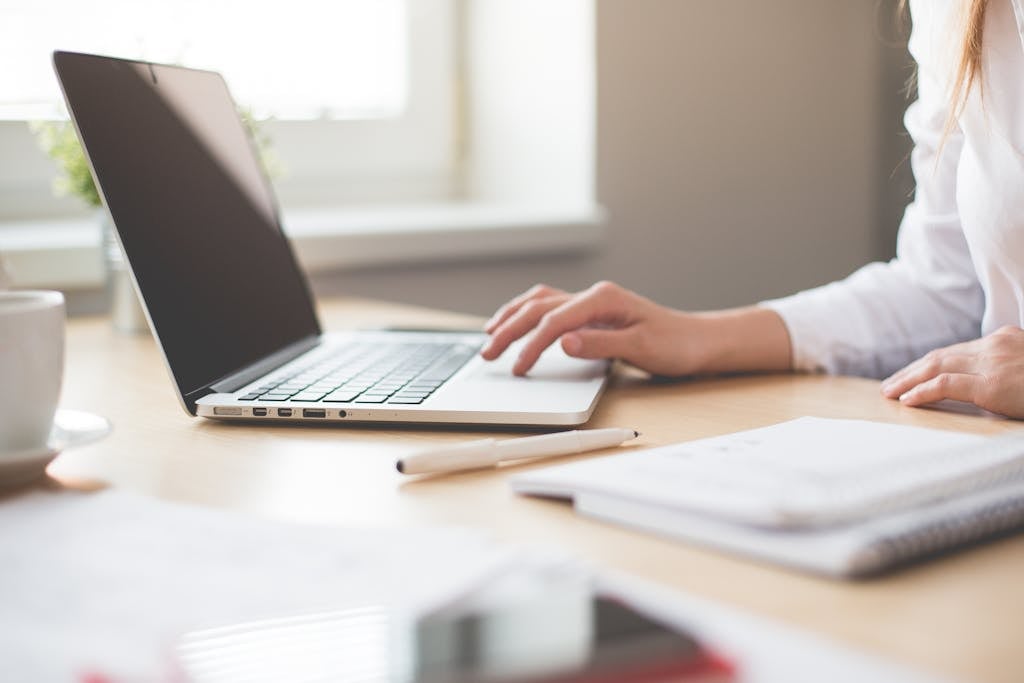 A businesswoman typing on a laptop at a bright indoor workspace with notebooks and a pen.