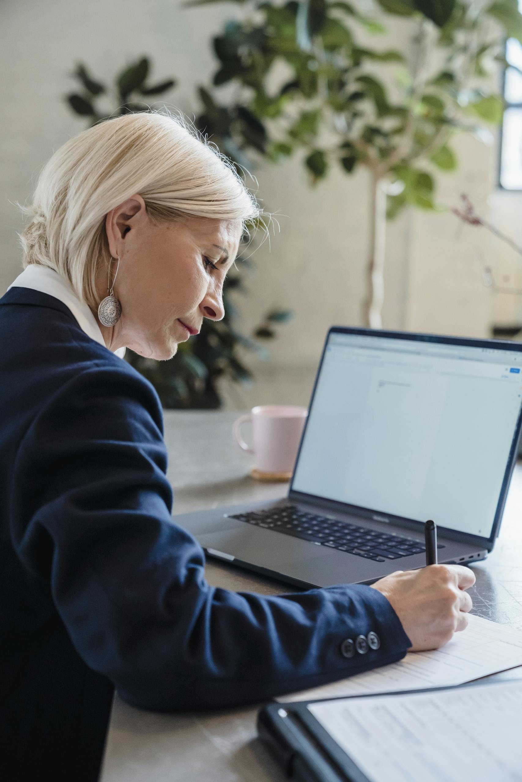 Elderly woman working on a laptop at office desk, writing notes with focus.