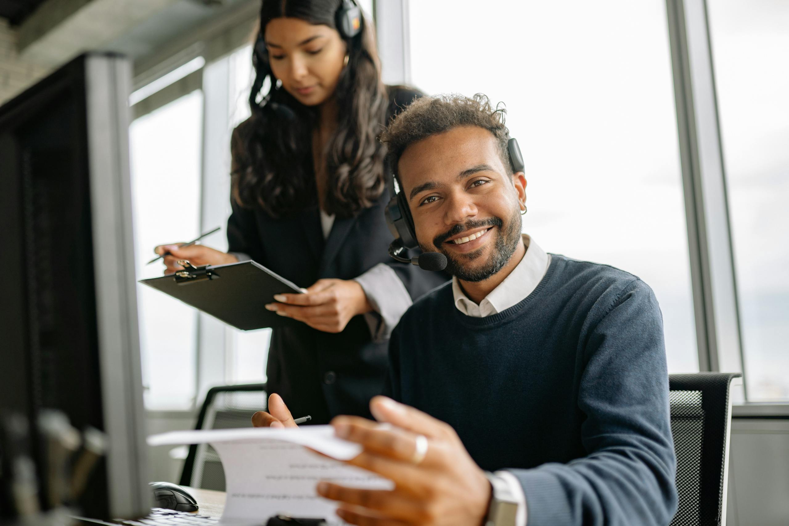 Friendly call center agent wearing a headset, talking and smiling at his desk with a colleague.